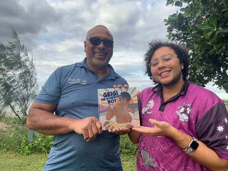 A Torres Strait Islander male smiling holding a children's book alongside a younger Torres Straight islander male wearing glasses and smiling.