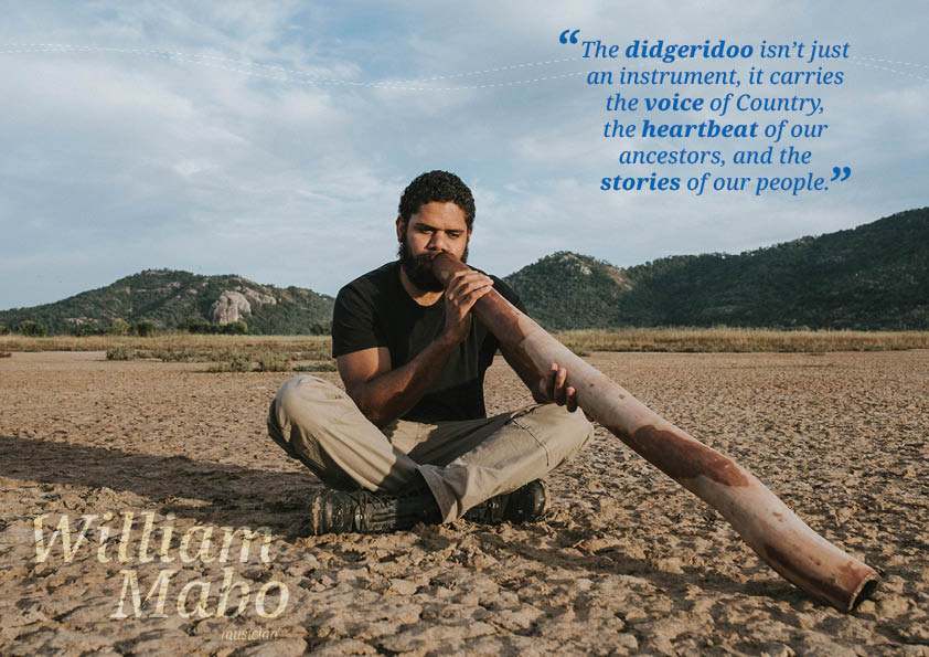 A First Nations man sits cross legged on a sandy ground playing a didgeridoo.