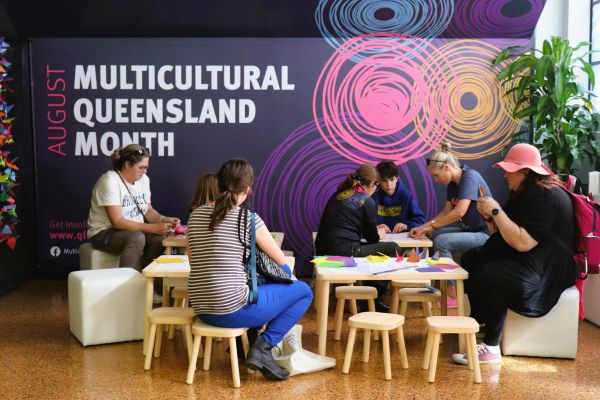 A group of people gathered around a table folding origami in front of a wall that says "August Multicultural Queensland Month".