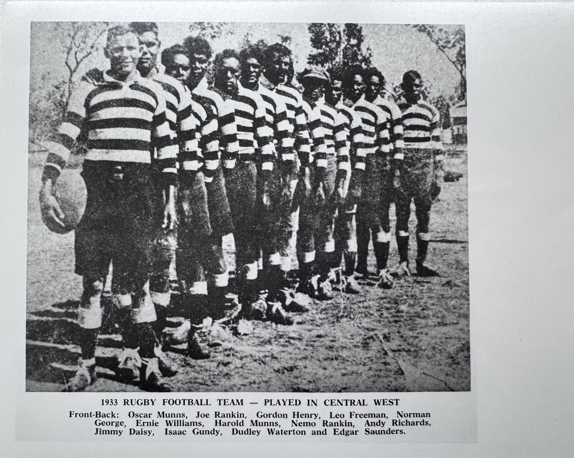 Black and white photo of First Nations male football players standing in a line dressed in striped football jerseys.